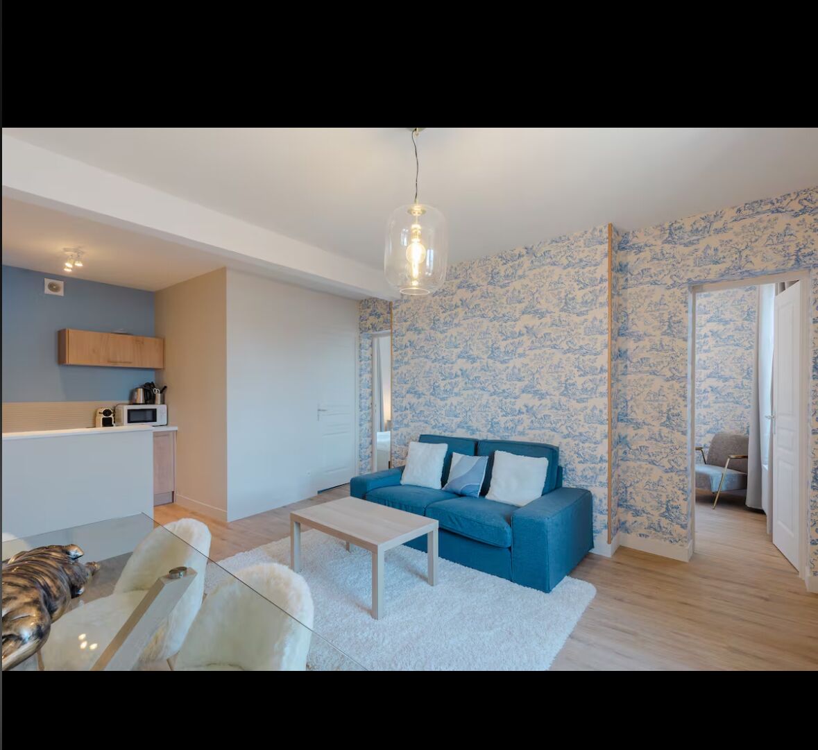 Ein modernes Wohnzimmer mit einem blauen Sofa, einem Glastisch und einem weißen Teppich. Es gibt eine Küchenzeile mit einer Mikrowelle und Holzschränken. Die Wände sind mit blumigem blauen Tapeten versehen.