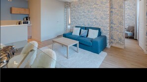 Sala de estar moderna com um sofá azul, uma mesa de centro de vidro e um tapete branco. Há uma área de cozinha com micro-ondas e armários de madeira. As paredes têm papel de parede floral azul.