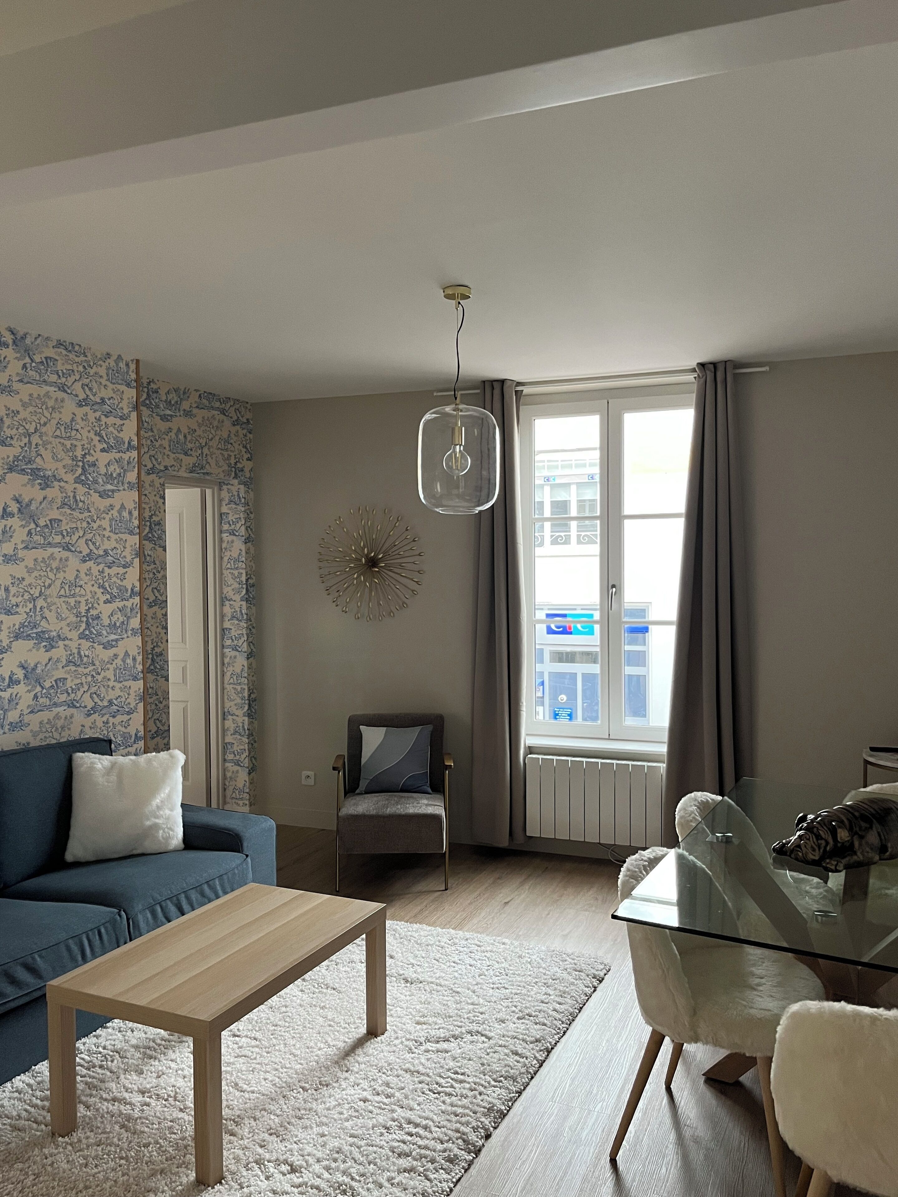 Un soggiorno con un divano blu, un tavolino in vetro, una poltrona, un elemento decorativo a parete e una finestra con tende.