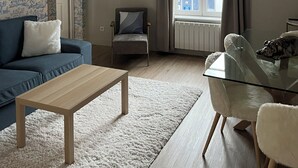 Uma sala de estar com um sofá azul, uma mesa de centro de vidro, uma cadeira, um objeto decorativo na parede e uma janela com cortinas.