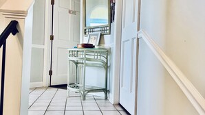 Un escalier recouvert de moquette mène à un palier qui donne sur une pièce avec une porte blanche, un miroir et une petite table sur laquelle se trouvent un bol et un cadre photo.