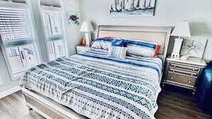 Une chambre à coucher avec un grand lit, une table de chevet, une lampe, un tableau encadré représentant des voiliers et une fenêtre avec des stores.