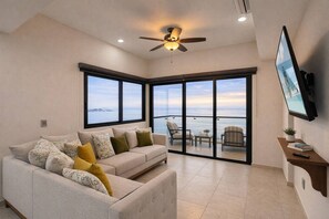 A modern living room with a sofa, a ceiling fan, a TV, and a balcony with a view. - Condo Premium - Malecón | Vista al mar | Grupos 6+ (Mazatlán)