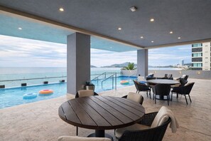 Área de piscina ao ar livre com vista para o mar, uma mesa redonda de madeira e cadeiras de descanso.