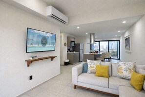 A modern living room with a sofa, a flat-screen TV mounted on the wall, and a kitchen area in the background. - Condo Premium - Malecón | Vista al mar | Grupos 6+ (Mazatlán)