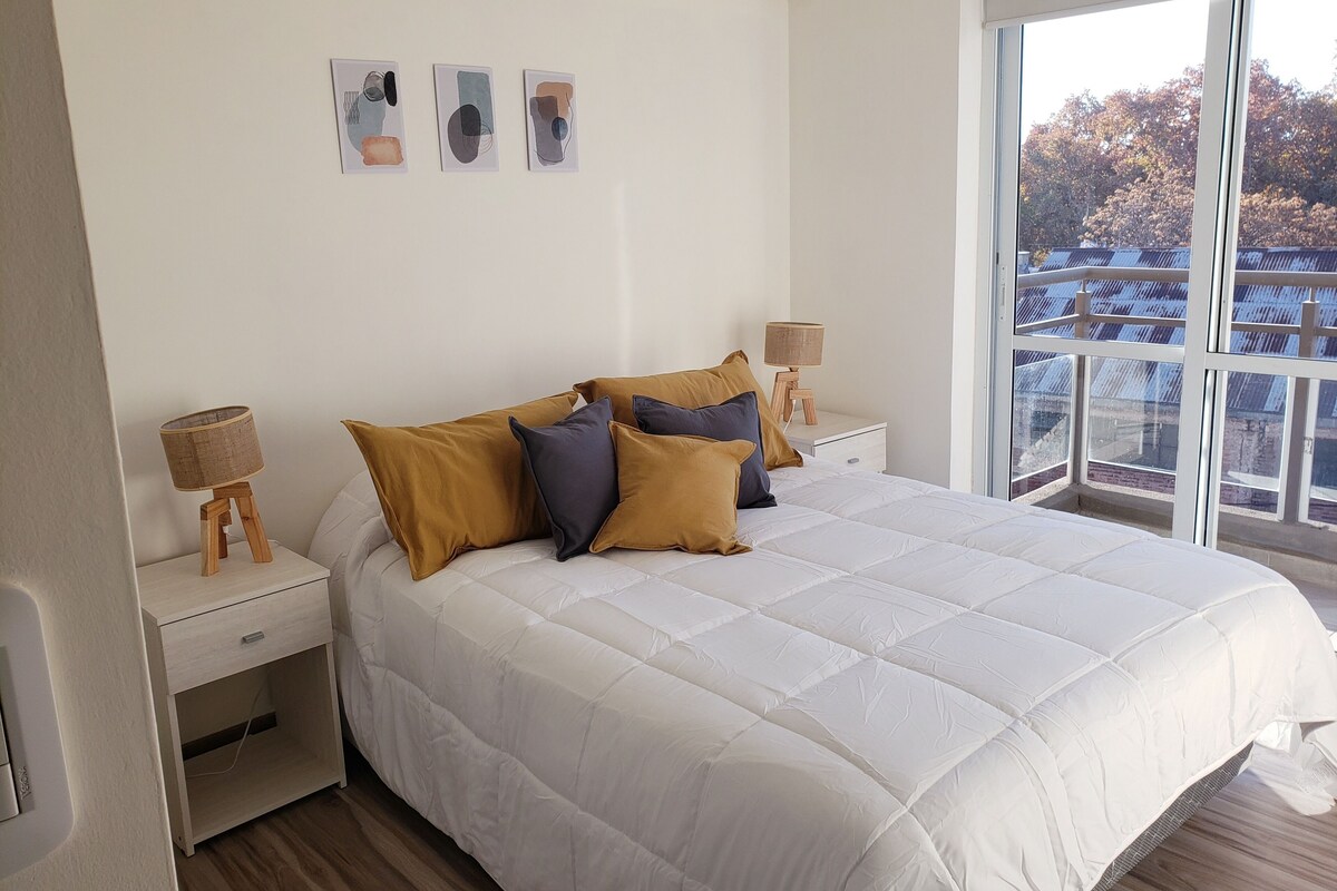 A bedroom with a large bed, two bedside tables, a window with a view, and three wall-mounted artworks.