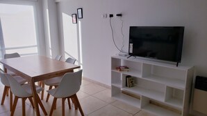 A modern living room with a wooden dining table, white chairs, a flat-screen TV on a white stand, and a tiled floor. - Patricias Park (Mendoza)