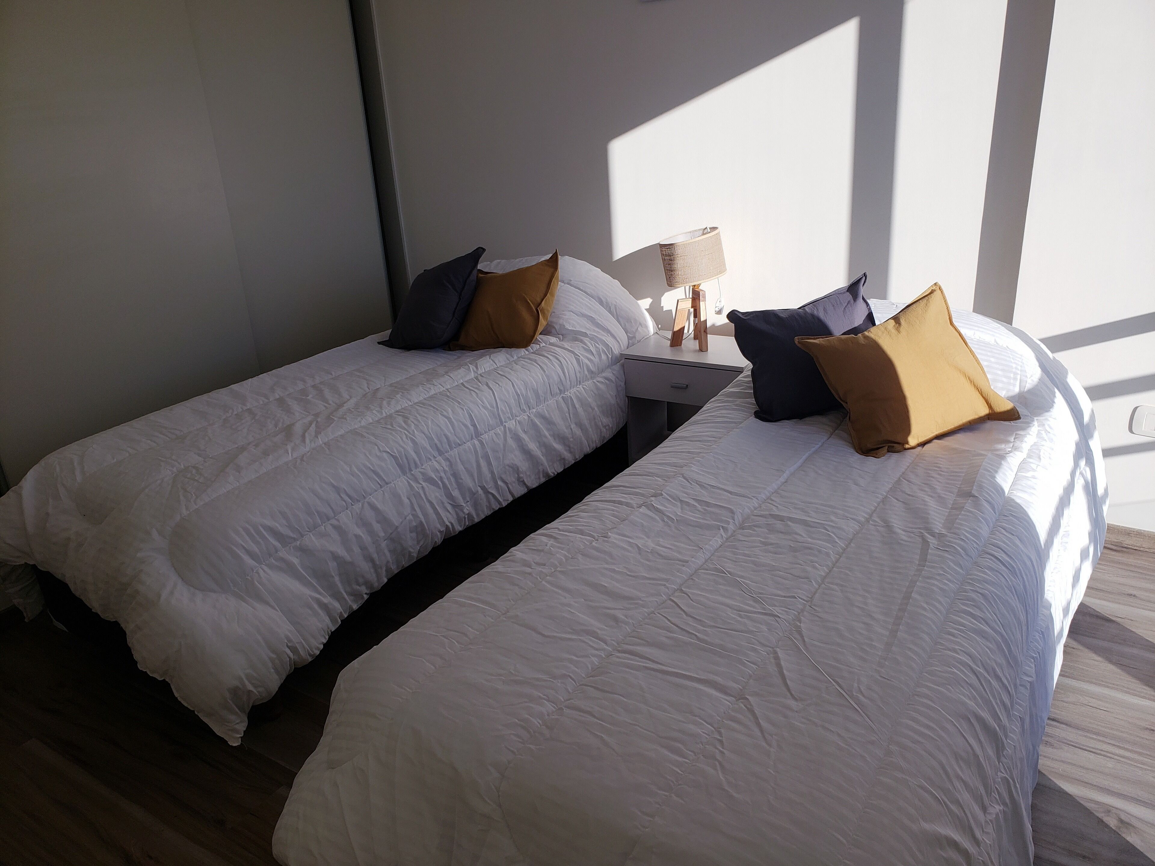 Habitación de hotel con dos camas individuales, una mesita de noche con lámpara y vistas al exterior.