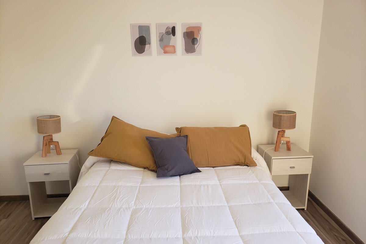 A neatly made bed with two pillows, a wooden nightstand with a lamp, and a wall-mounted picture above the bed.
