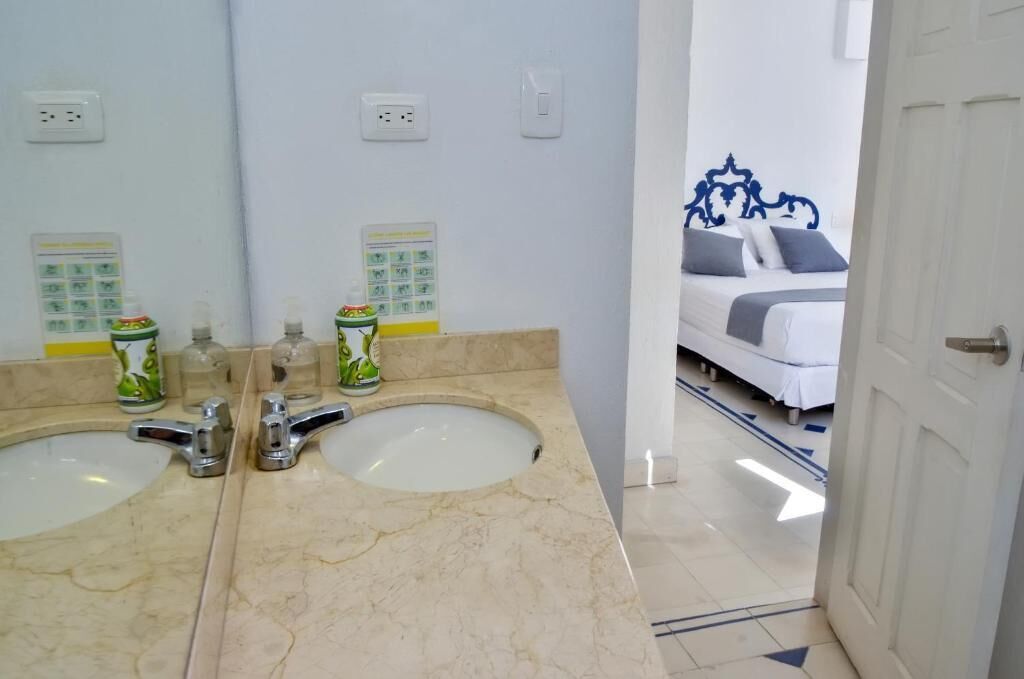 Un baño con dos lavamanos, un espejo y una cama visible a través de una puerta abierta.