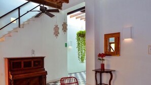 Una stanza con pavimento a scacchi, un pianoforte in legno, sedie rosse, un tavolino e una ventola a soffitto.