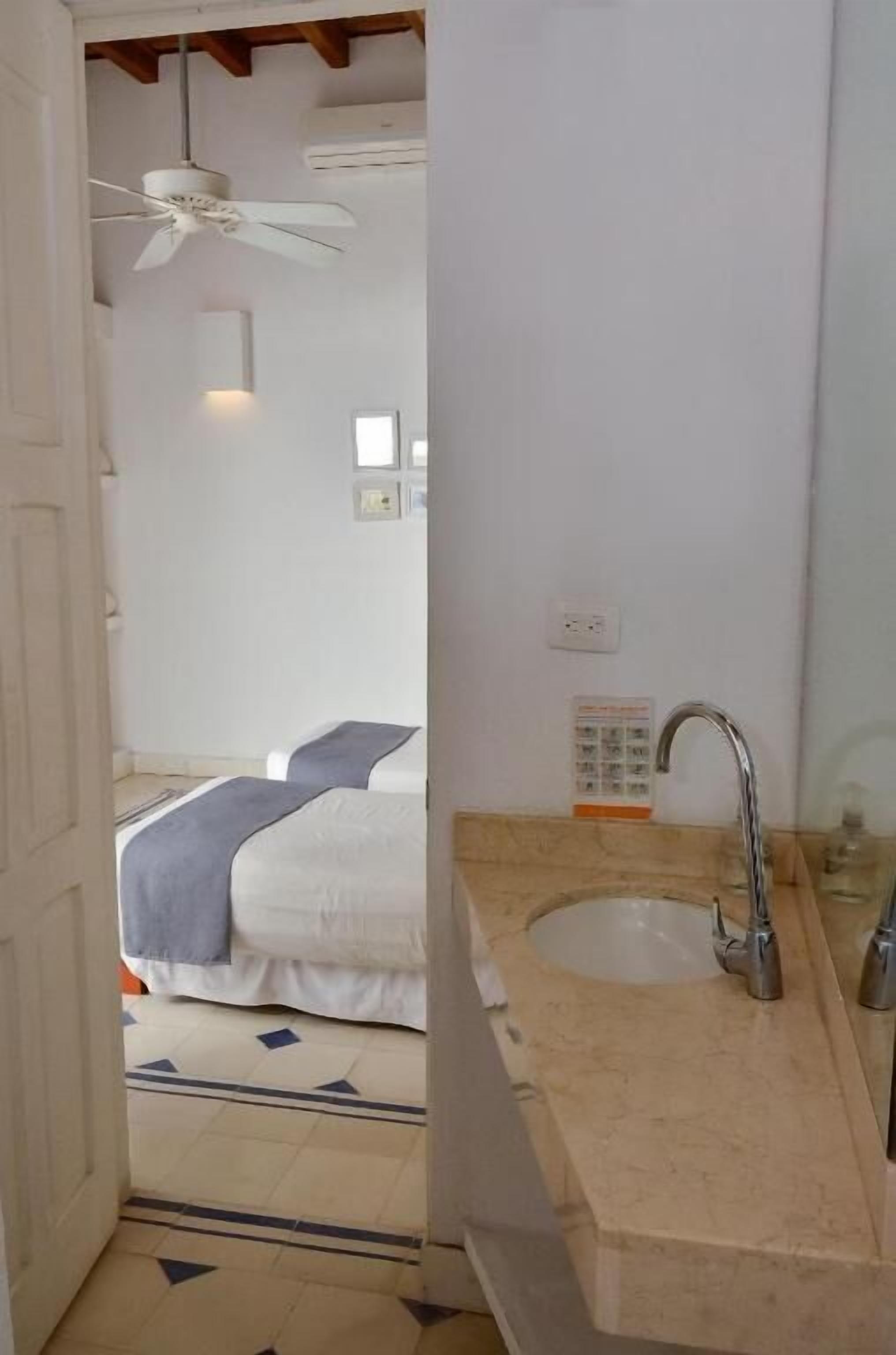 A hotel room with a bed, a ceiling fan, a bathroom with a sink and mirror, and a tiled floor.