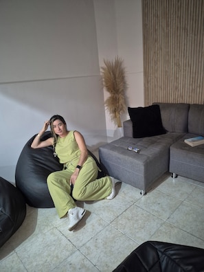 Una persona sentada en un sillón de tina negro en una sala de estar moderna con un sofá gris, un jarrón alto con plumas de pampa y un revestimiento de pared de listones de madera.
