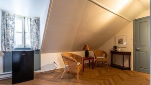 A cozy room with a herringbone wooden floor, two wicker chairs, a small table with a lamp, a black refrigerator, and a door with a framed picture.