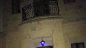 Un edificio de piedra con puerta de madera, un pequeño balcón con barandilla de hierro forjado y un farol iluminado en azul sobre la puerta.