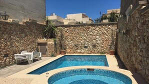 Una piscina rectangular con un jacuzzi en el centro, rodeada de muros de piedra y algunas plantas en macetas.
