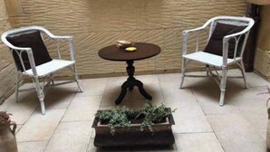 Un patio con dos sillas blancas de mimbre, una mesita redonda y una jardinera con plantas verdes.