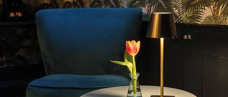 A room with a blue chair, a small round table with a lamp, and a vase with a single tulip.