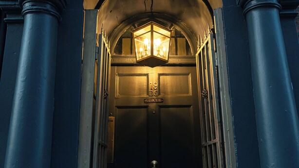 Ingresso di un edificio blu scuro, con una lanterna accesa sopra la porta, colonne ai lati e un muro in mattoni sullo sfondo.