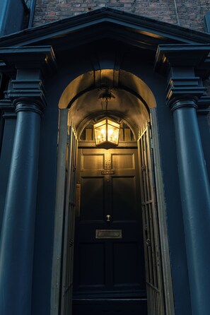 Entrada de un edificio azul oscuro con un farol iluminado sobre la puerta, columnas a ambos lados y un muro de ladrillo al fondo.