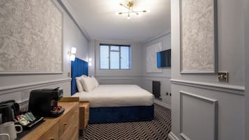 A bedroom with a bed, bedside table, coffee maker, and a window with blinds.