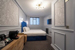 A bedroom with a bed, bedside table, coffee maker, and a window with blinds.