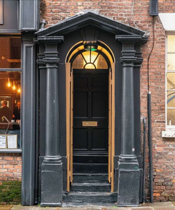 A black door with a small window above it, flanked by two columns, and a brick wall in the background.