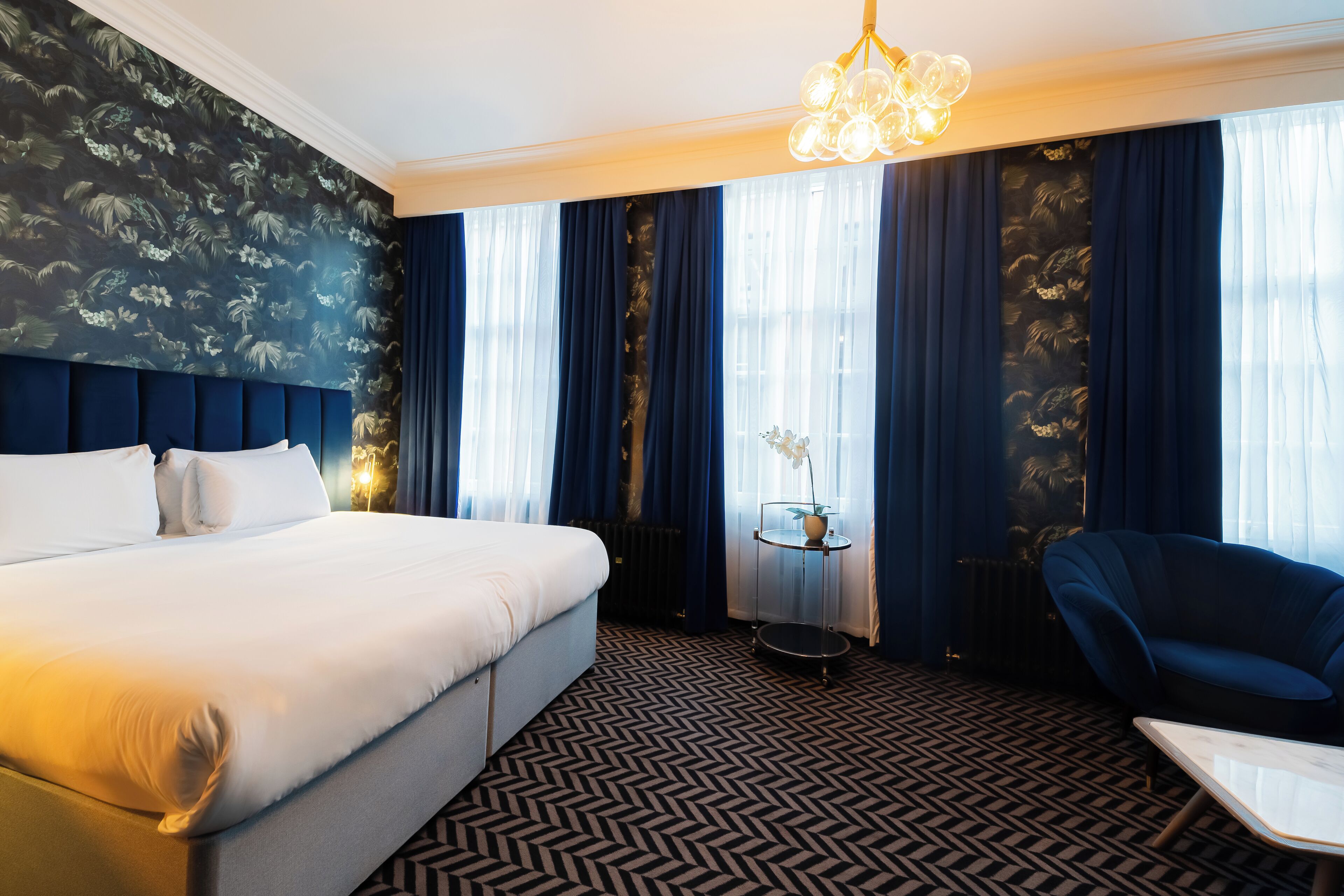 A hotel room with a large bed, a round table with a vase, a blue armchair, and a patterned carpet.