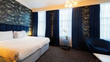 A hotel room with a large bed, a round table with a vase, a blue armchair, and a patterned carpet.