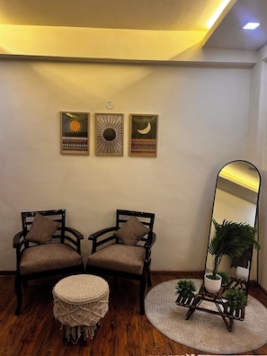 Una habitación con dos sillas de madera oscura con cojines, un taburete redondo tejido, un espejo con una planta y tres cuadros en la pared.