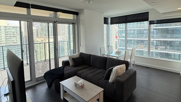 Ein modernes Wohnzimmer mit Ecksofa, Couchtisch und Fernseher. Große Fenster bieten Blick auf eine Stadtlandschaft.
