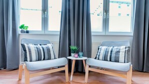 Une pièce comprenant deux fauteuils gris à revêtement en tissu, dotés de coussins à rayures, une petite table ronde ornée d’une plante et de deux tasses, et une fenêtre avec des rideaux gris.