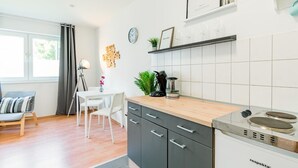 Une cuisine moderne avec un plan de travail en bois, des armoires noires et une cuisinière encastrée. On y trouve un coin repas avec une table et des chaises blanches, ainsi qu’un canapé gris. Une horloge et un tableau encadré sont accrochés au mur.