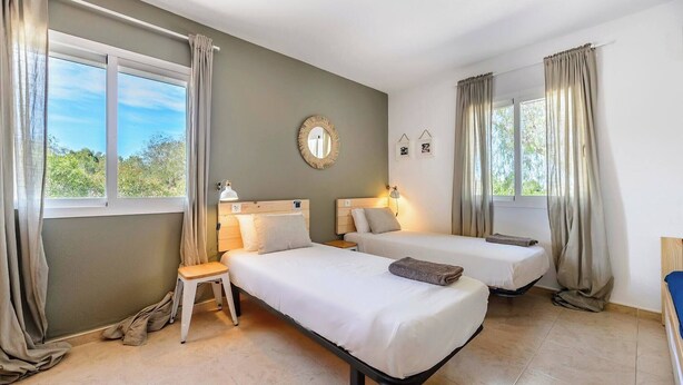 Ein Hotelzimmer mit zwei Betten, einem kleinen Tisch, einem Stuhl, einem runden Spiegel und Blick auf Grünflächen.