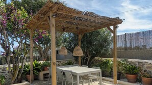 Eine überdachte Terrasse mit Tisch und Stühlen, umgeben von Topfpflanzen und einer Pergola.