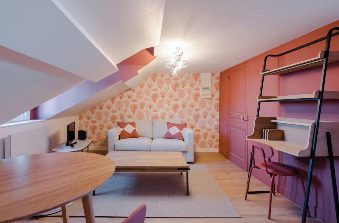Un espacio habitable compacto con sofá, mesa de centro y pequeña mesa de comedor. Una estantería escalonada está apoyada en la pared, y el fondo está cubierto con un papel tapiz estampado.