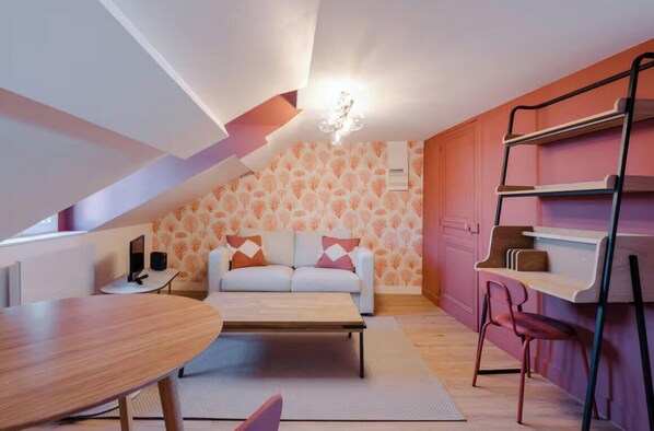 A compact living space with a sofa, coffee table, and a small dining table. A ladder shelf is against the wall, and there is a patterned wallpaper on the back wall. - Charming renovated two rooms wifi (Cabourg)