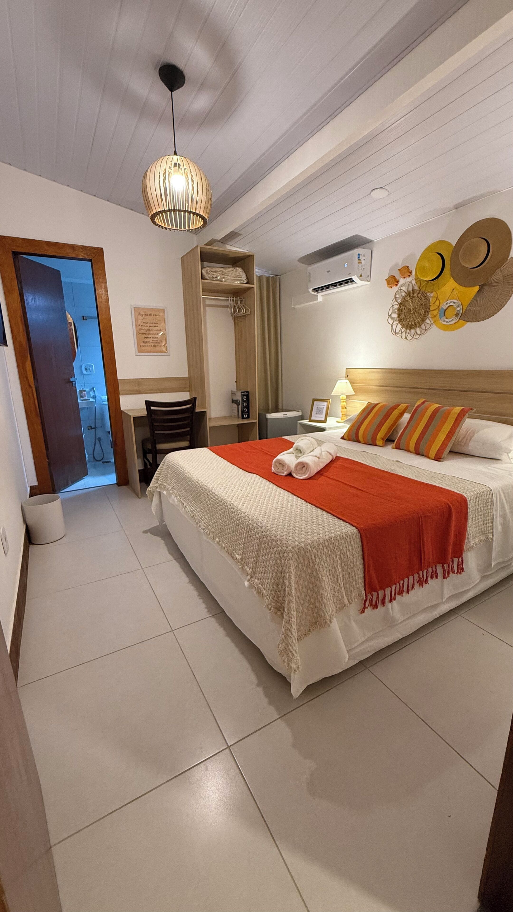 Quarto de hotel com uma cama grande, cabeceira de madeira, mesa de cabeceira com abajur, ar condicionado fixado na parede e porta para o banheiro.