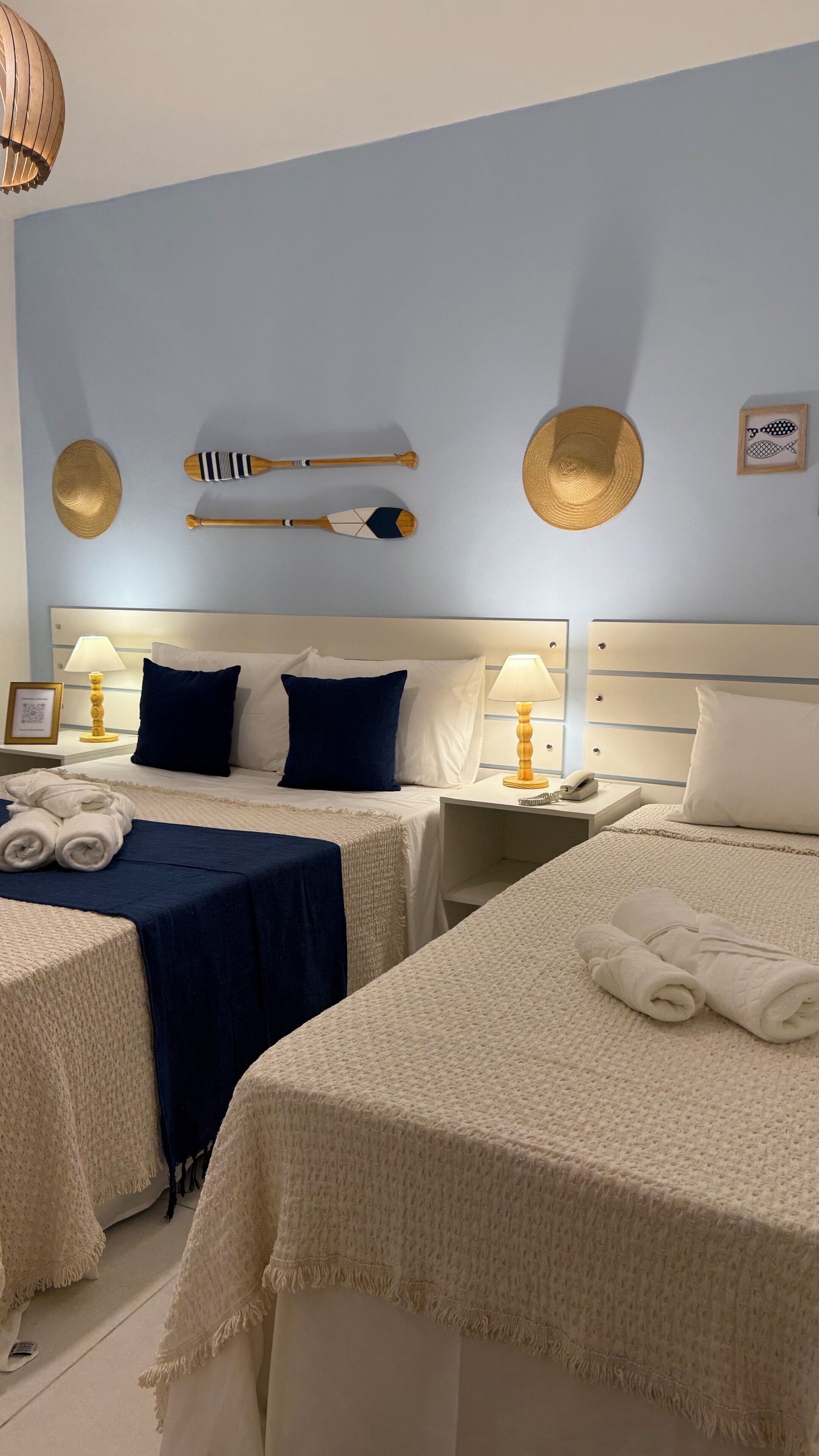 A hotel room with two beds, each with a white bedspread and blue pillows. There are two bedside tables with lamps, a framed picture, and a small decorative item on the wall.