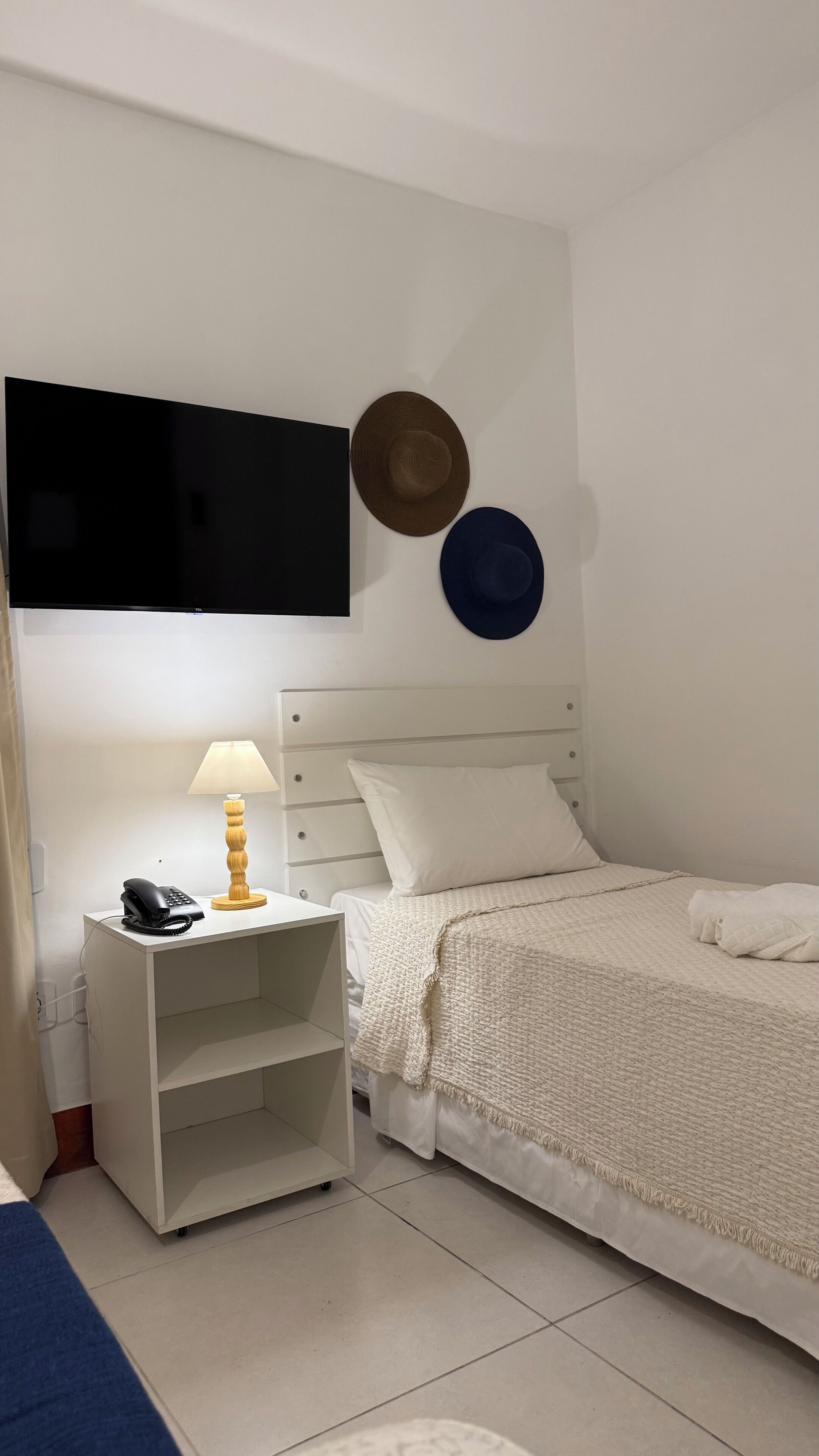 A bedroom with a bed, a bedside table with a lamp, a phone, and two hats on the wall.