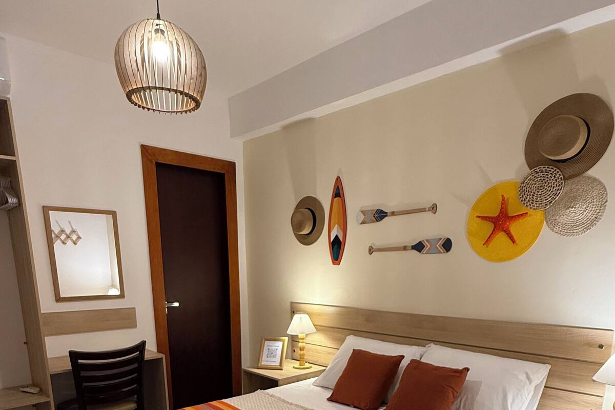 Een netjes ingerichte hotelkamer met een groot bed, een bureau en een stoel. Er is een wandplank met decoratieve objecten en een plafondventilator.