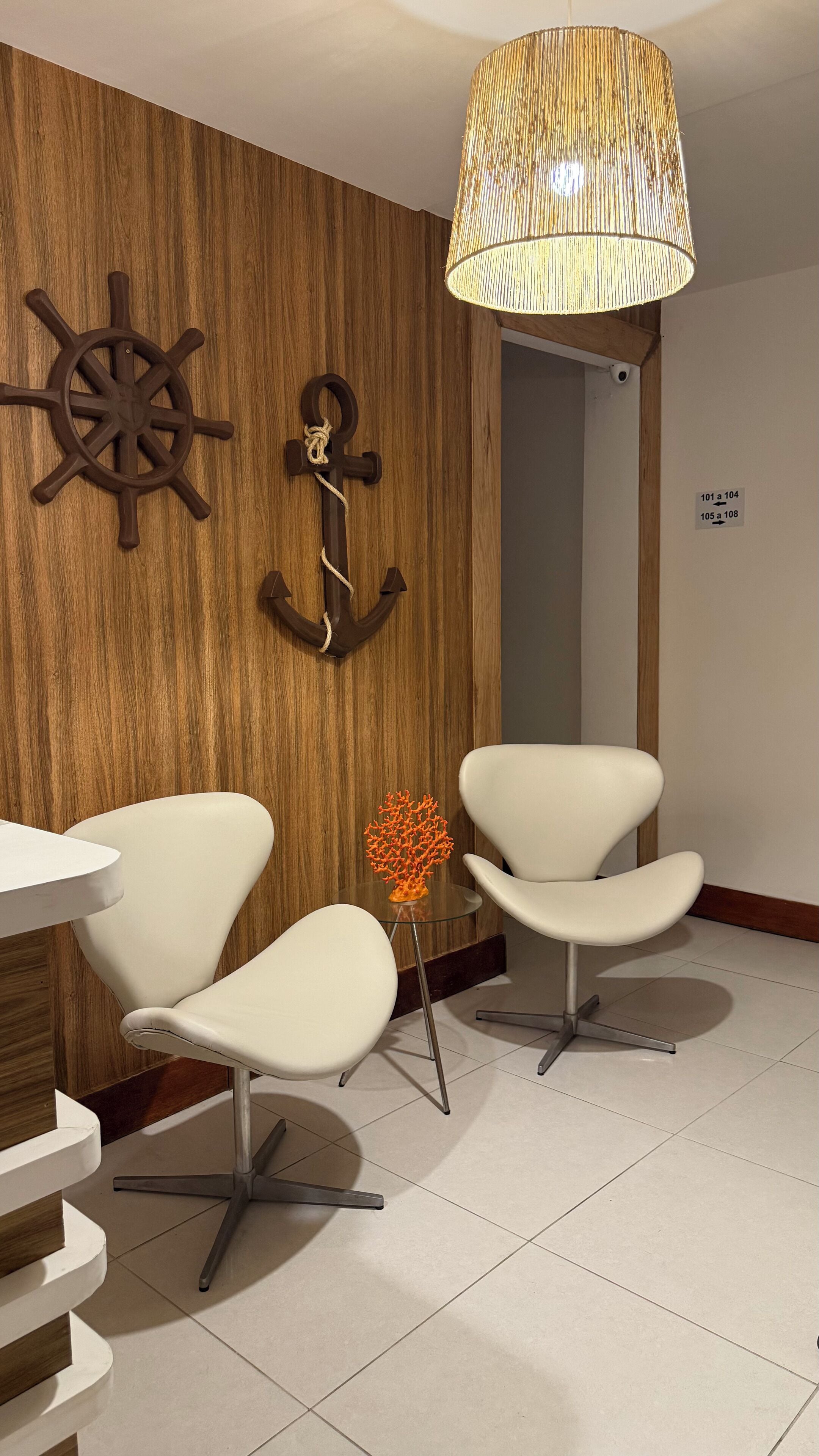 A modern room with two white chairs, a wooden wall with a ship's wheel and anchor decoration, and a hanging lamp.