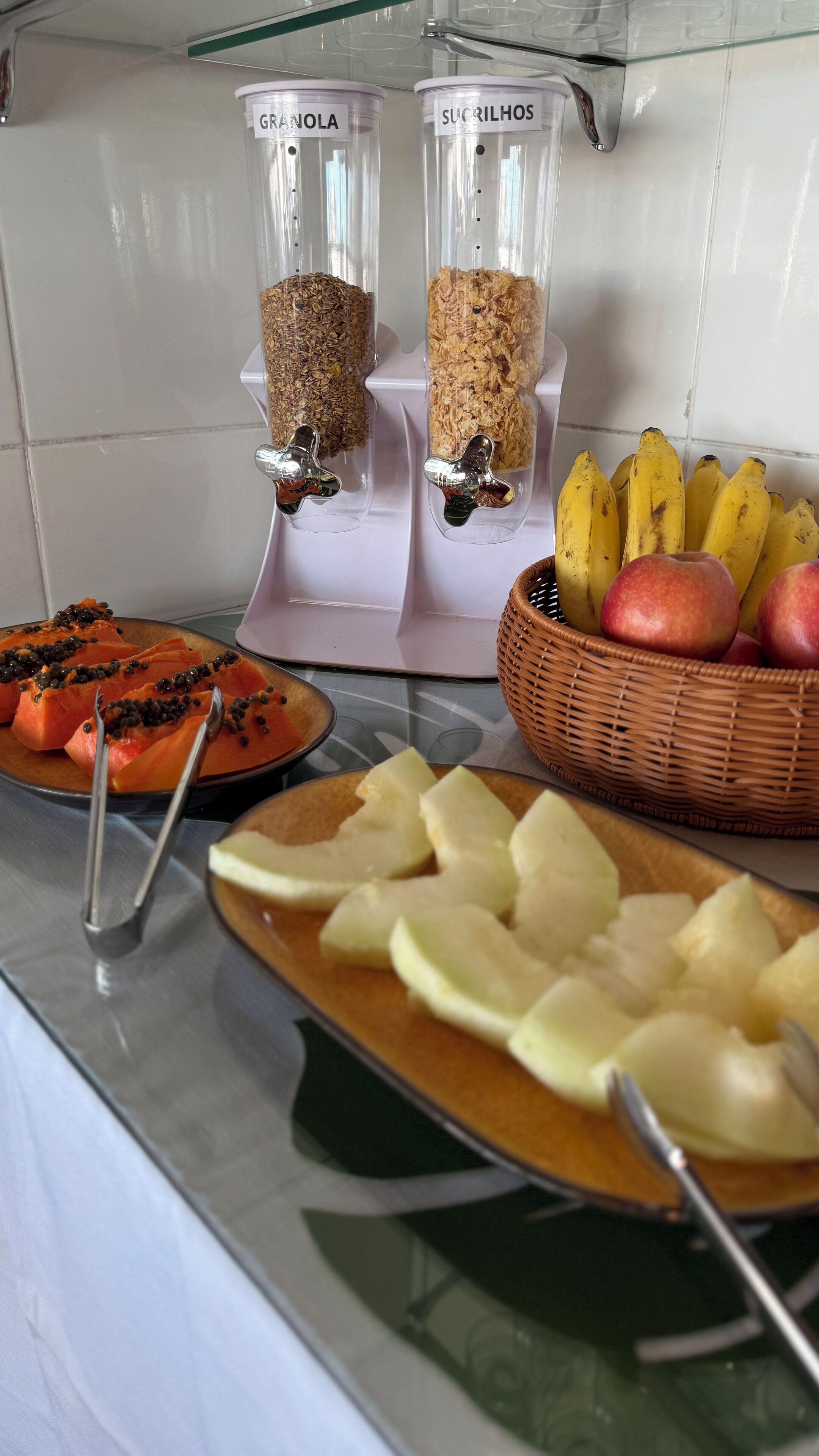 A kitchen counter with food items, including sliced apples, a bowl of bananas, and a plate of sliced papaya with black seeds. There are also two containers labeled "GRANOLA" and "SUCRILHOS".