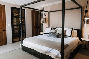 Uma cama com dossel em tons neutros, uma pequena mesa redonda e um lustre.