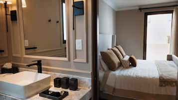 A hotel room with a large mirror, a white sink, a marble countertop, a bed with multiple pillows, and a window with curtains.