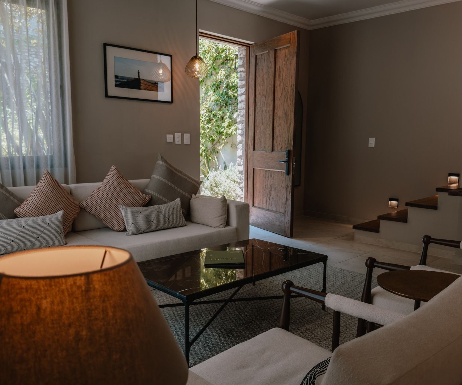 A modern living room with a sofa, coffee table, and chairs. There is a window with curtains, a picture on the wall, and a door leading outside.