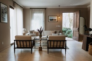 Una sala de estar moderna con un sofá, dos sillones, una mesa de centro y una mesita auxiliar con una lámpara. Hay un ventanal con cortinas translúcidas, un cuadro enmarcado en la pared y una puerta que da al exterior.