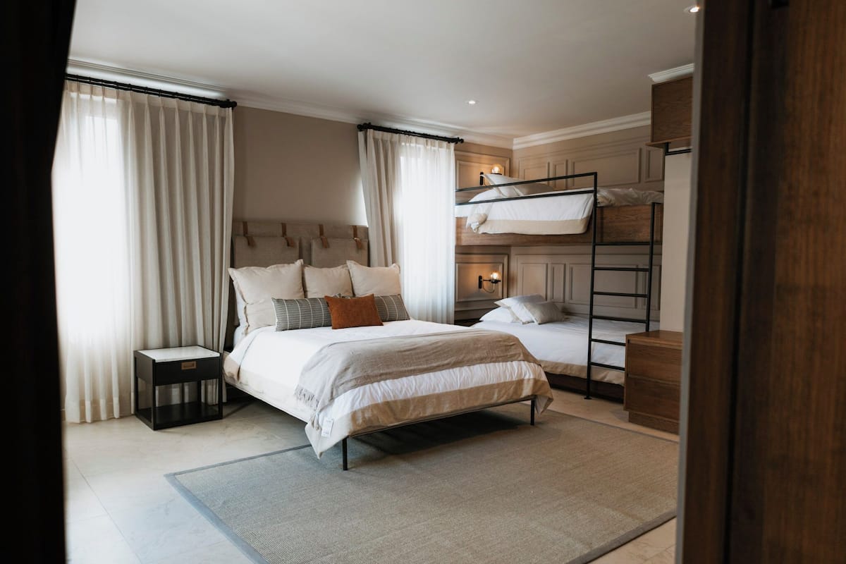 A bedroom with a bunk bed, a bedside table, a nightstand, and a large window with curtains.