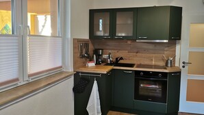 Une cuisine de taille réduite, avec des meubles de couleur vert foncé, un four encastré et une fenêtre équipée de stores.