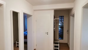 Un couloir doté d’un plancher en bois, de murs blancs et d’une porte donnant sur une chambre à coucher, où se trouvent un lit et une fenêtre.
